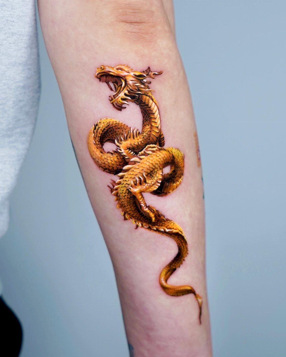 Golden Chinese dragon tattoo coiled on a person's forearm against a grey background