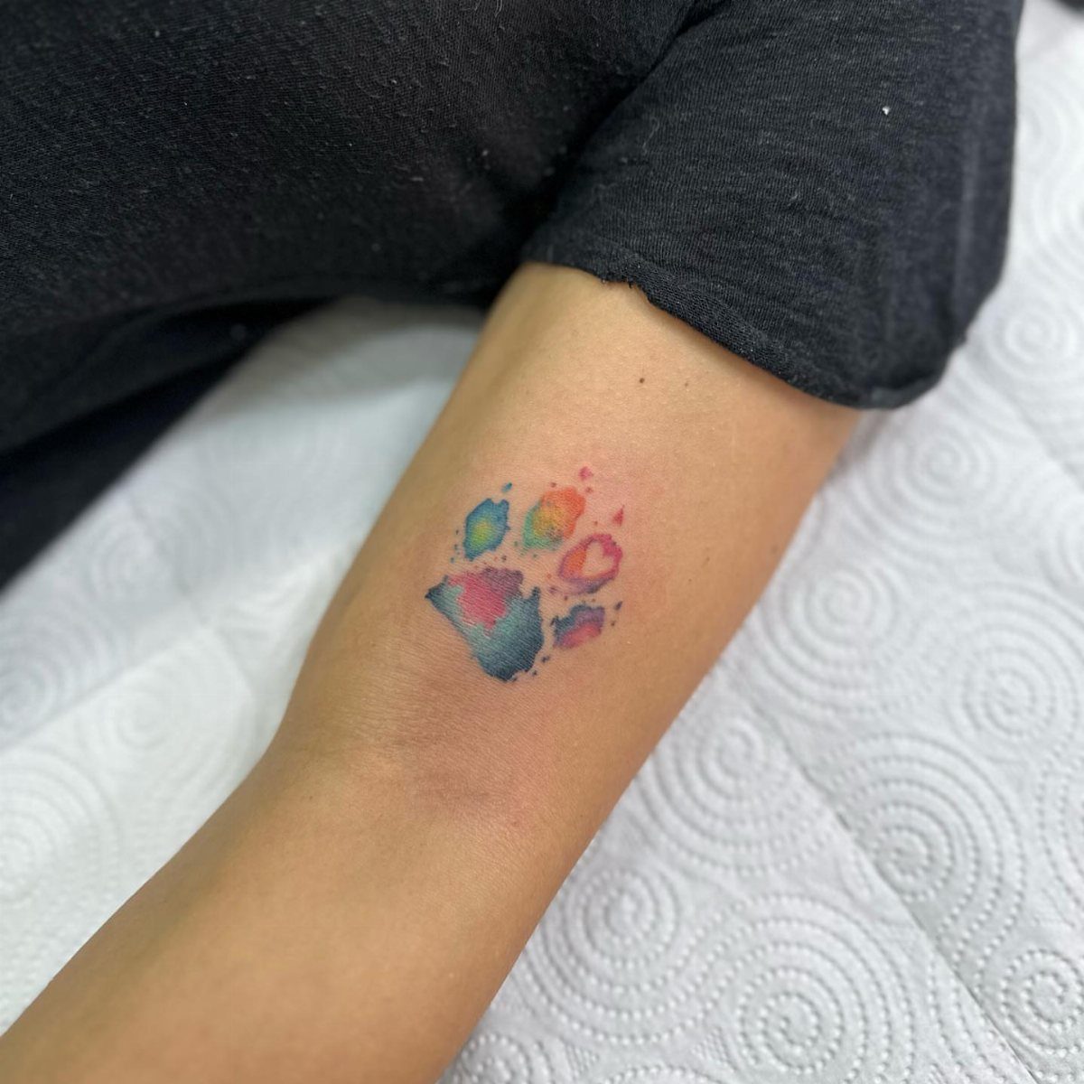 Watercolor dog paw tattoo with vibrant blue, pink, and yellow colors on a forearm.