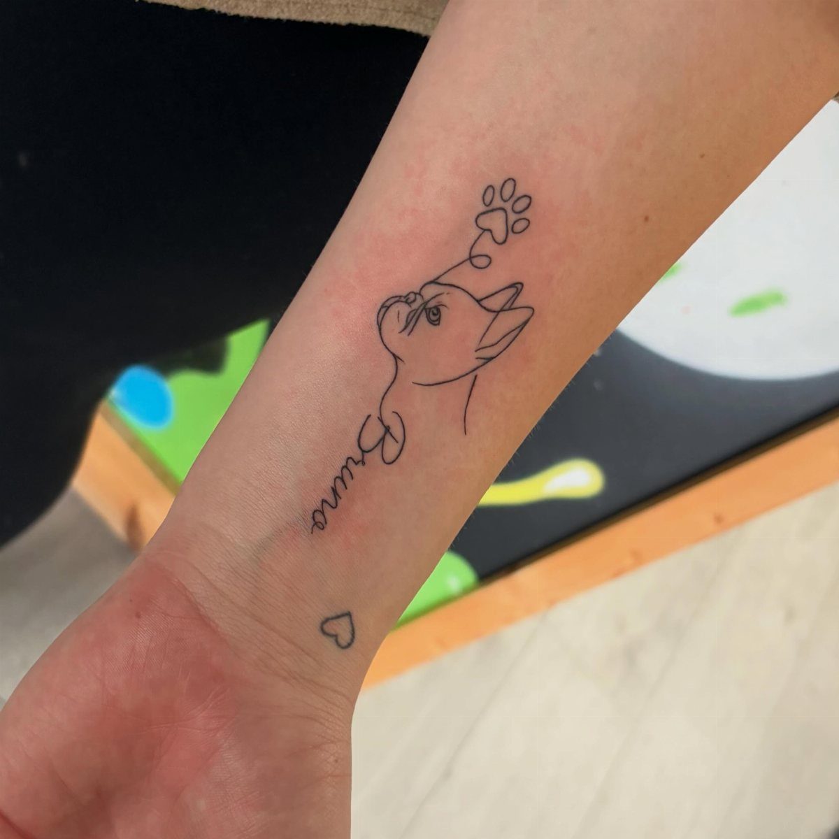 Forearm tattoo featuring a minimalist dog profile, the name Bruno, and a paw print.