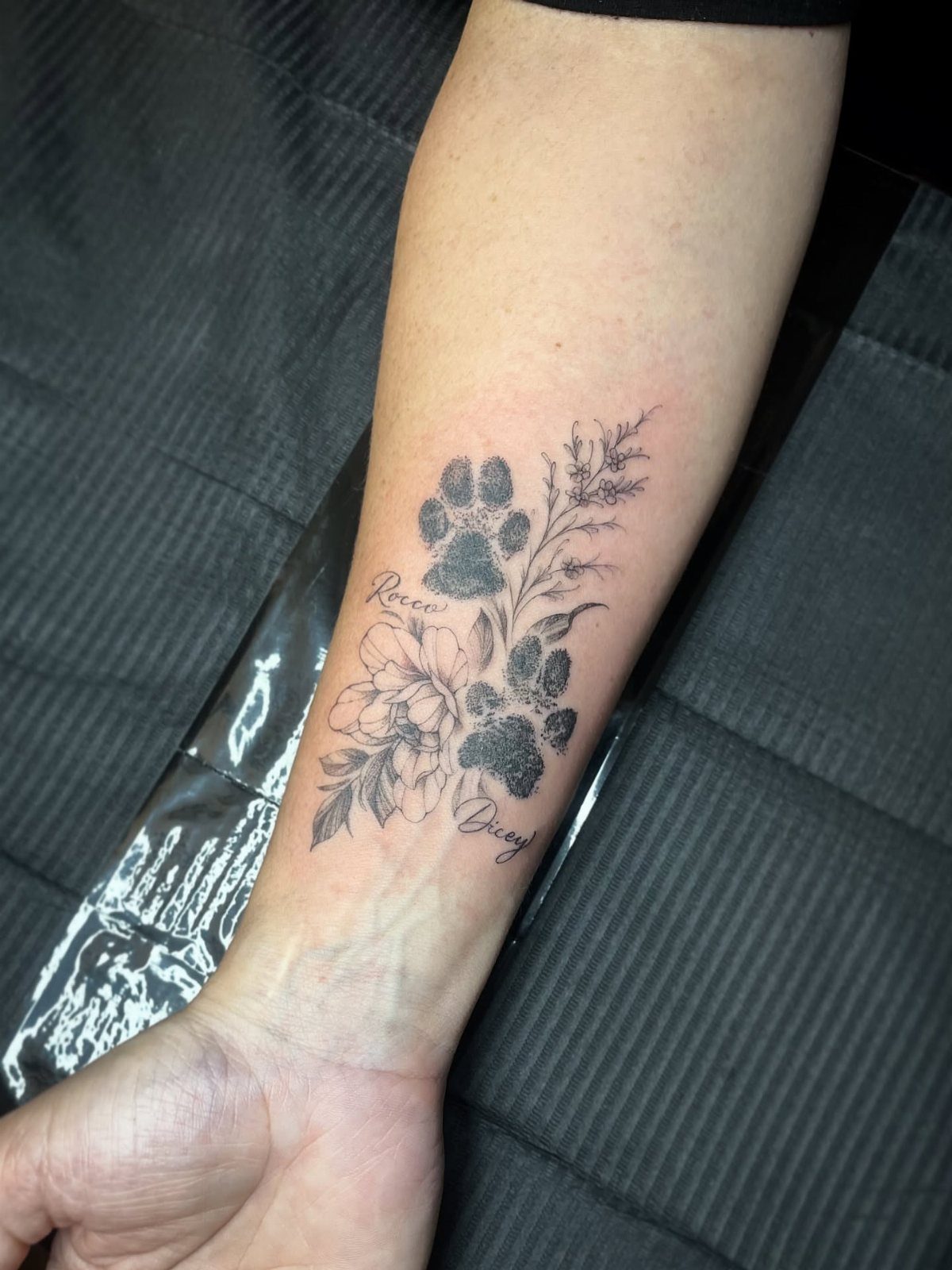 Dog paw print tattoo on a forearm with flowers and memorial names.