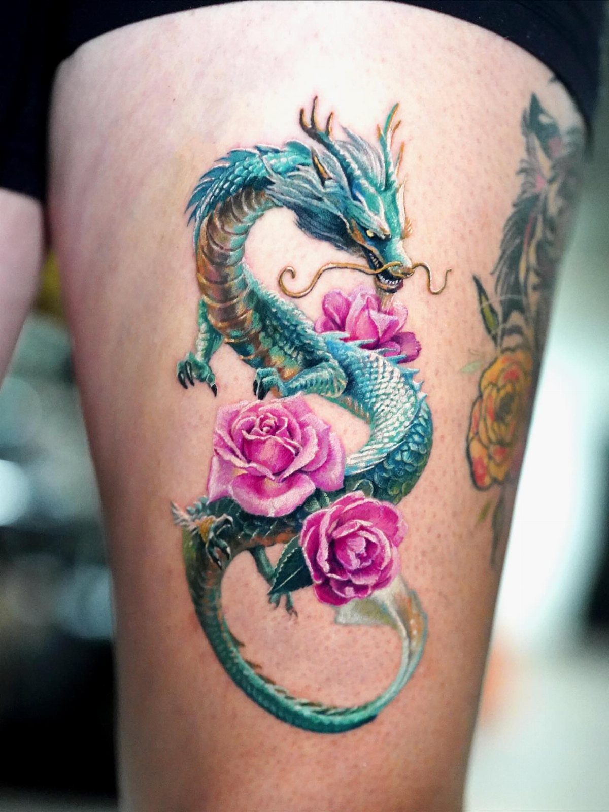 Teal and gold dragon tattoo entwined with pink roses on a person's thigh