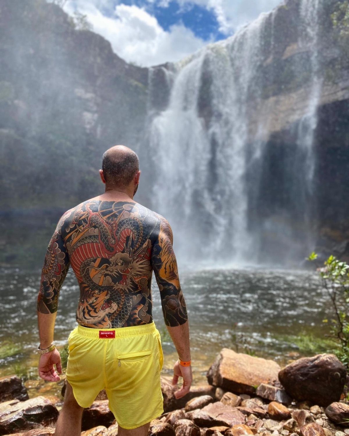 Man with a large red dragon tattoo on his back facing a waterfall