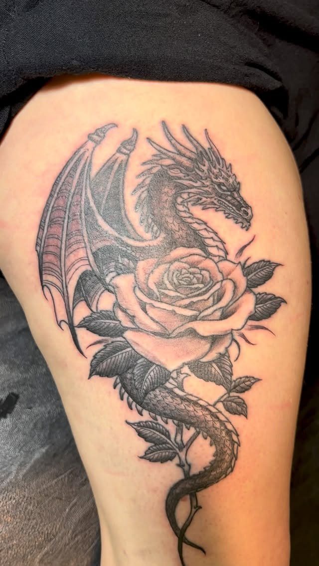 Dragon and rose tattoo in black and grey ink on a person's thigh.
