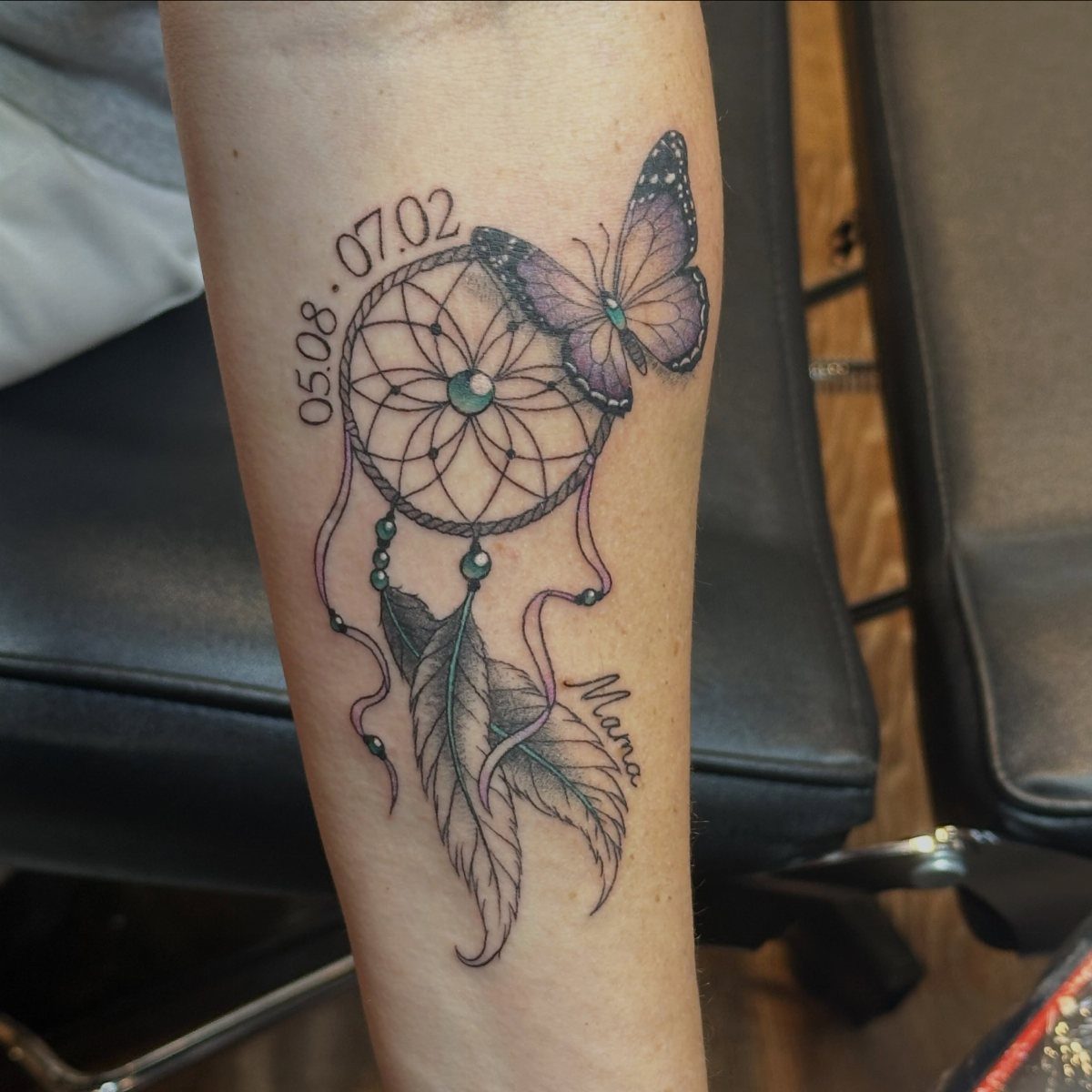 Dreamcatcher tattoo with a purple butterfly, feathers, and dates on a forearm.