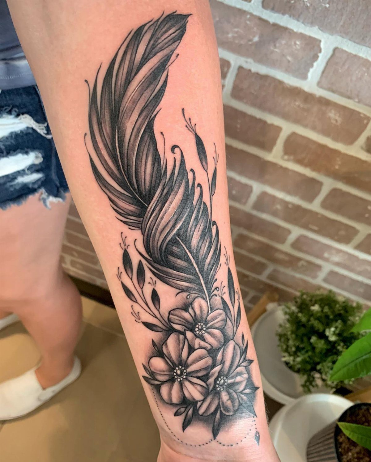 Detailed 3D feather tattoo with flowers on a person's forearm against brick wall