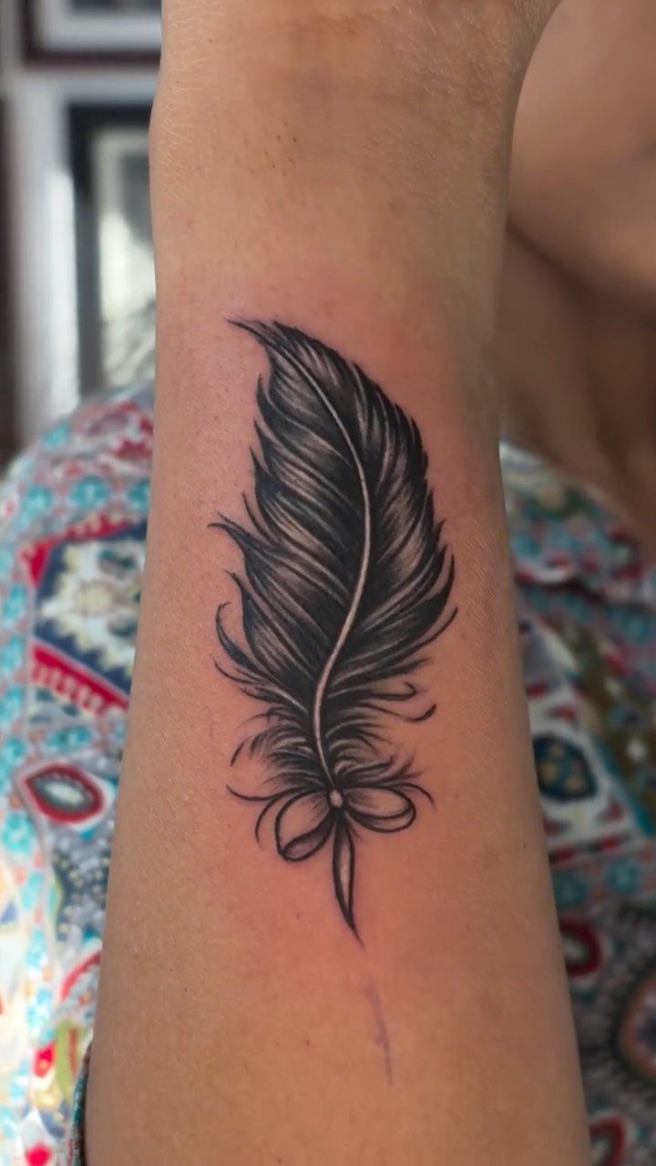 Black ink feather tattoo with a bow on a person's forearm