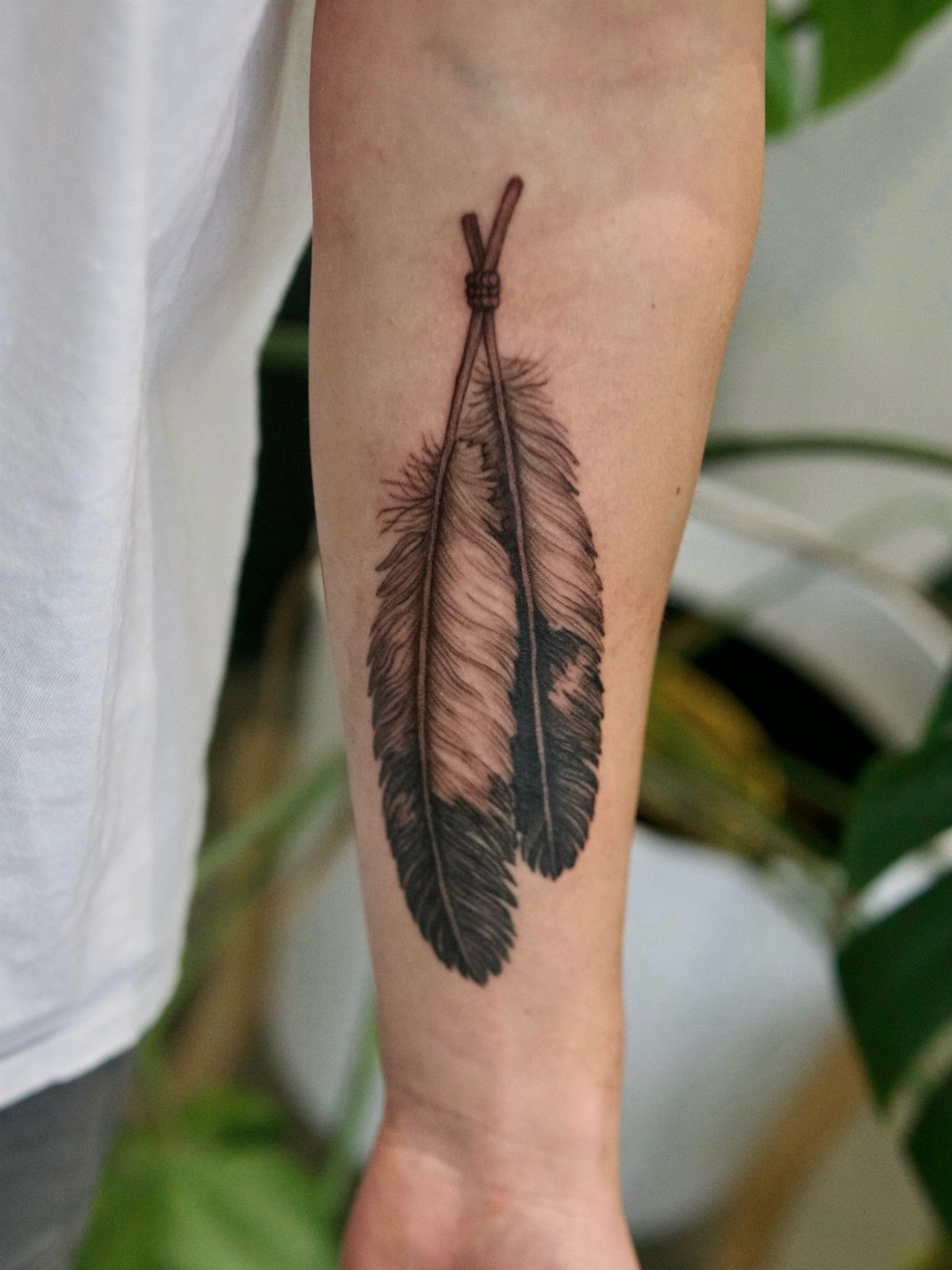 Two feathers tied together in a black and grey tattoo on a forearm