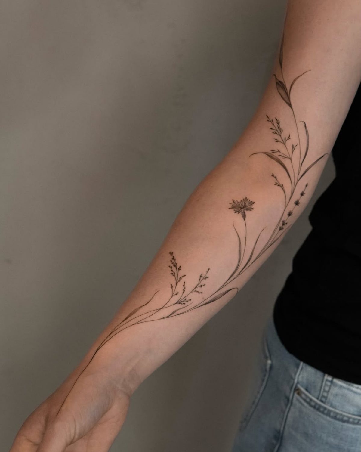 Fine line wildflower tattoo extending along a person's forearm from wrist to elbow.