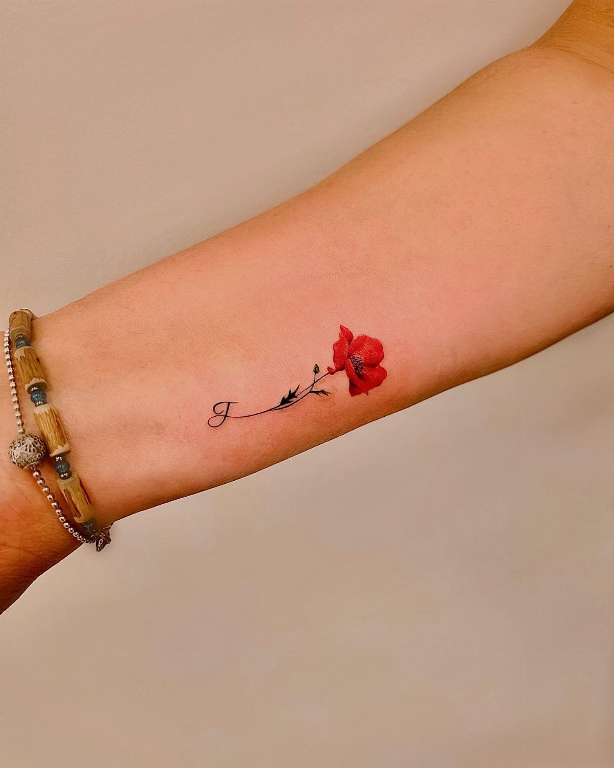 Red poppy flower tattoo with a cursive letter J on a forearm