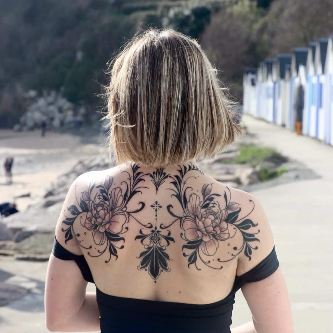 Symmetrical black ink floral tattoo across the upper back and shoulders of a woman