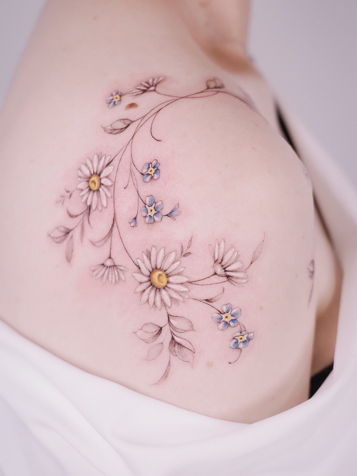 Daisy flower tattoo with small blue blossoms and leaves on a person's shoulder