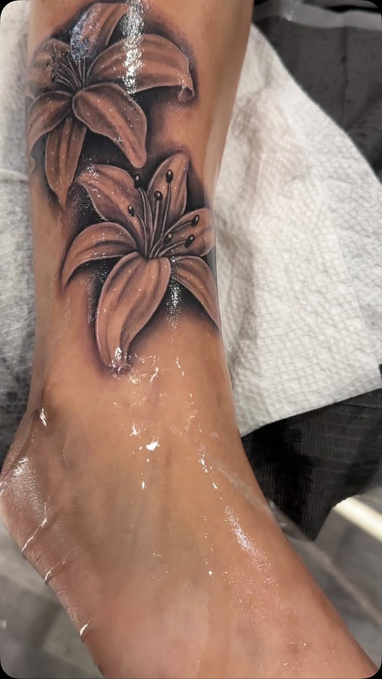 Two realistic lily flower tattoos in black and grey ink on an ankle.