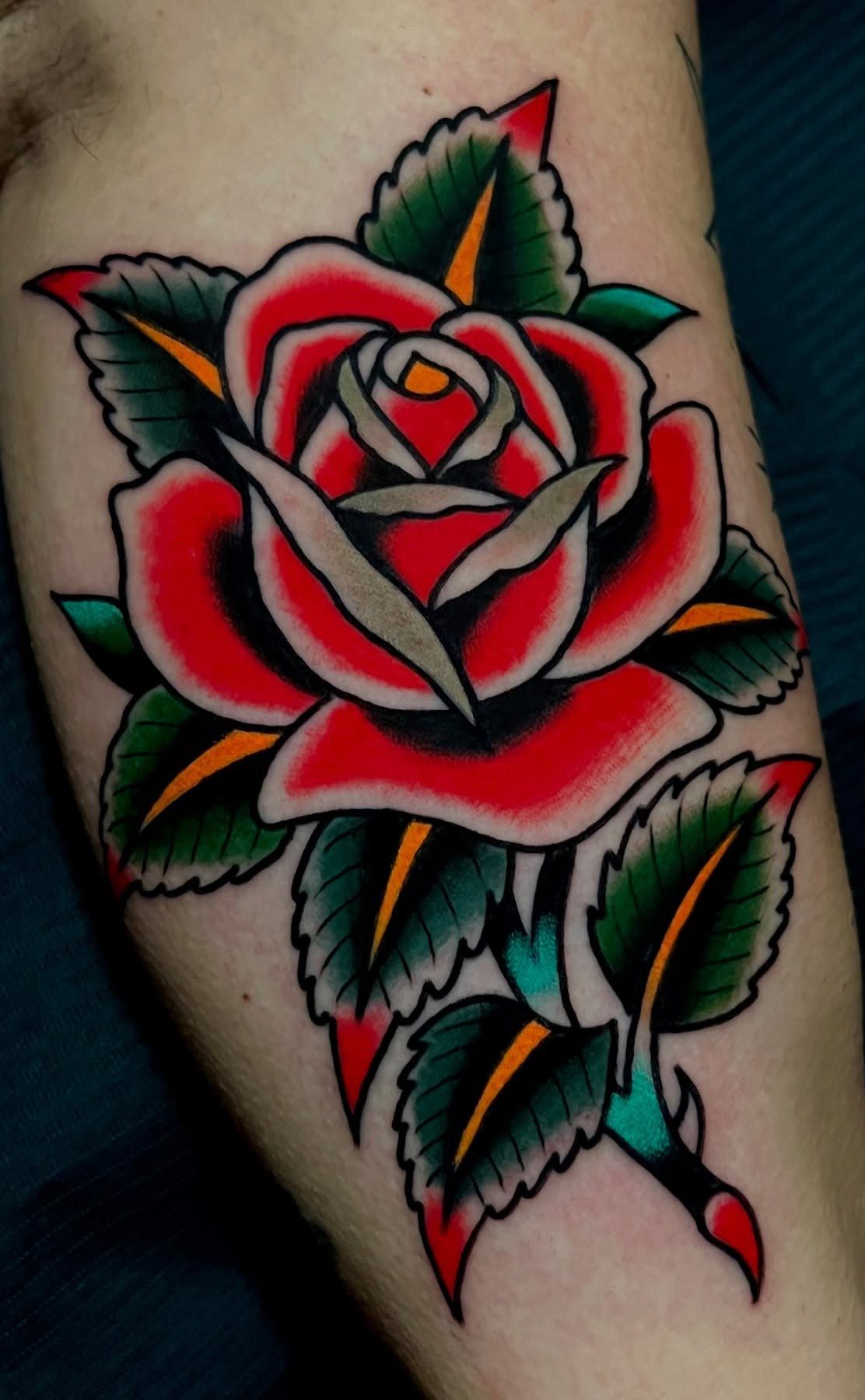 Red rose tattoo with bold black outlines and green leaves on skin.