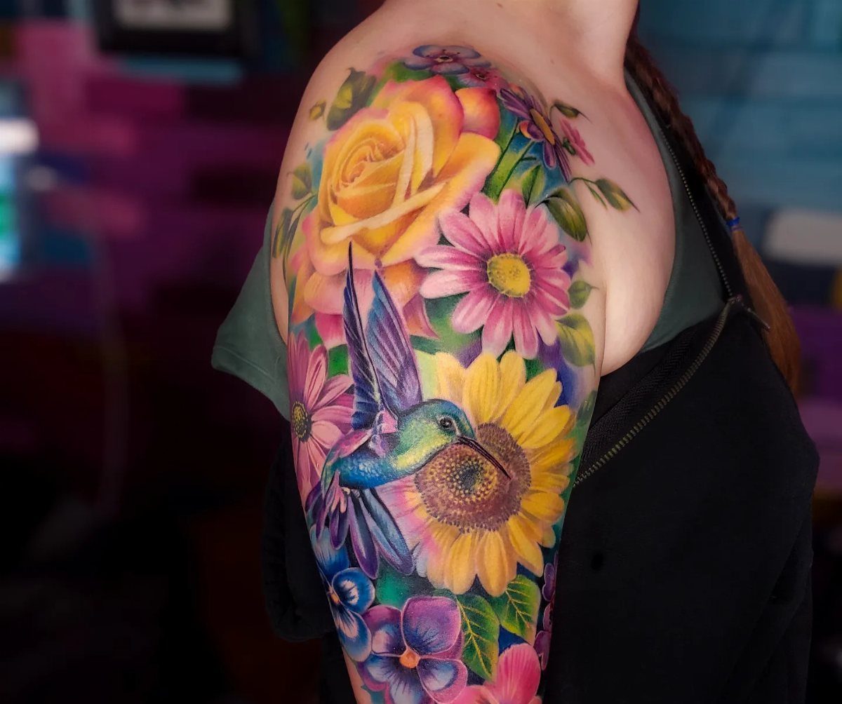 Colorful floral tattoo on a shoulder featuring a hummingbird, rose, and sunflower.