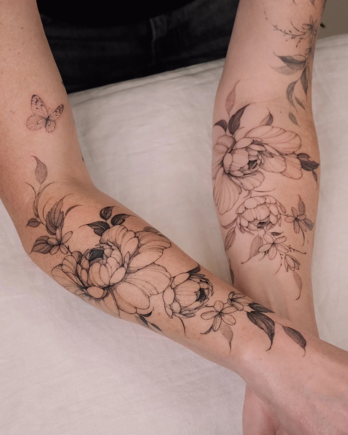Black ink peony tattoos with leaves and a butterfly on two forearms.