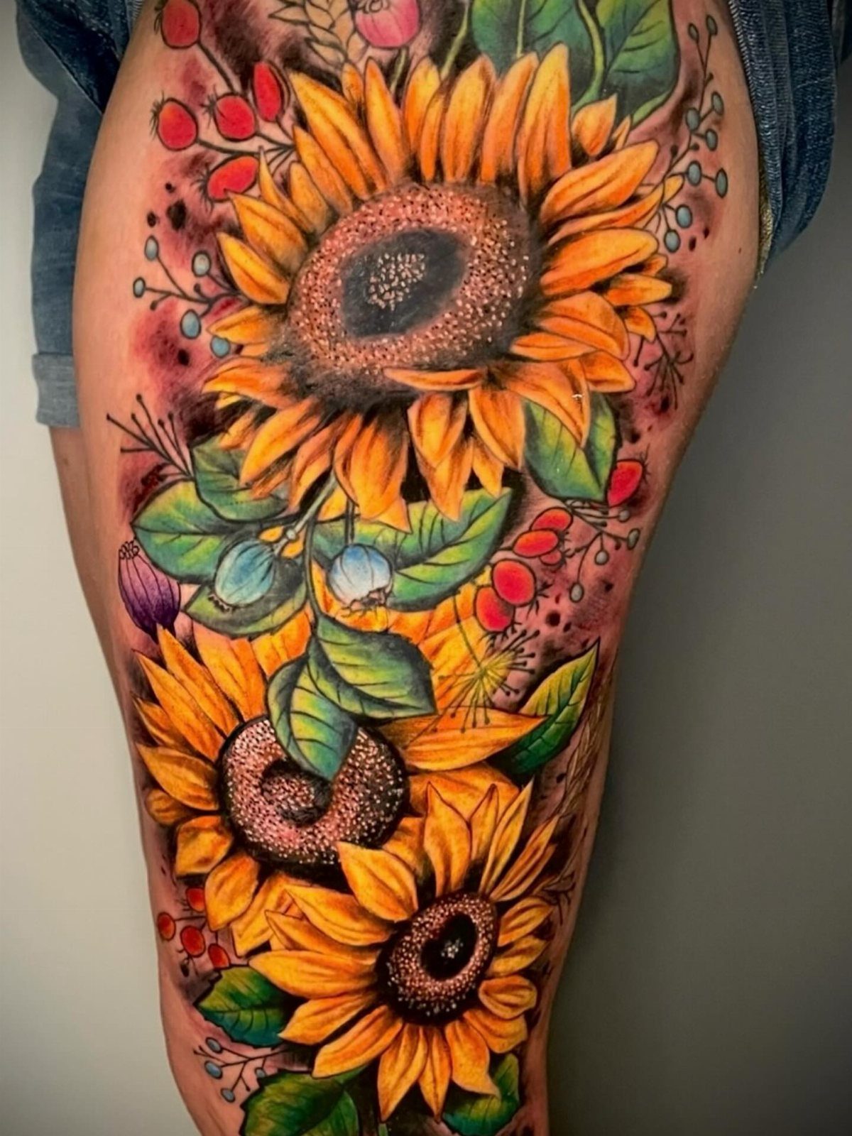 Colorful thigh tattoo featuring three large yellow sunflowers with berries and green leaves.
