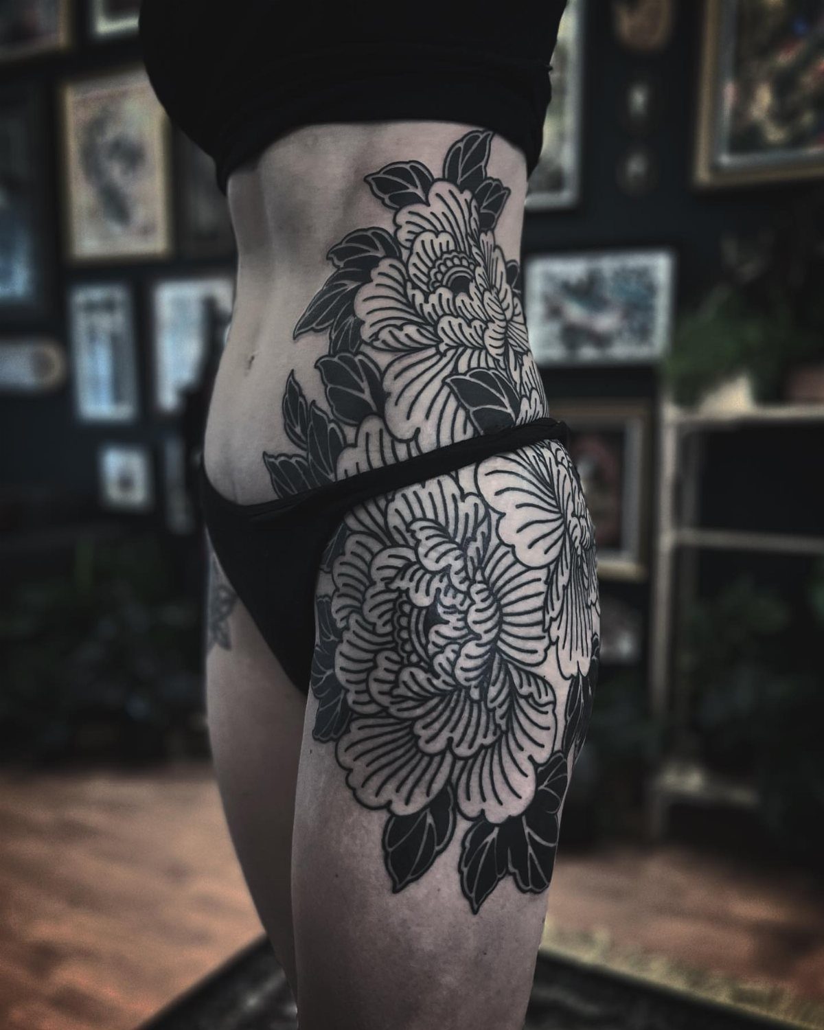 Large black ink peony tattoos with overlapping petals on a person's thigh and hip.
