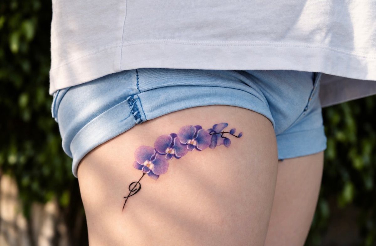 Purple orchid tattoo on a person's upper thigh wearing blue denim shorts.
