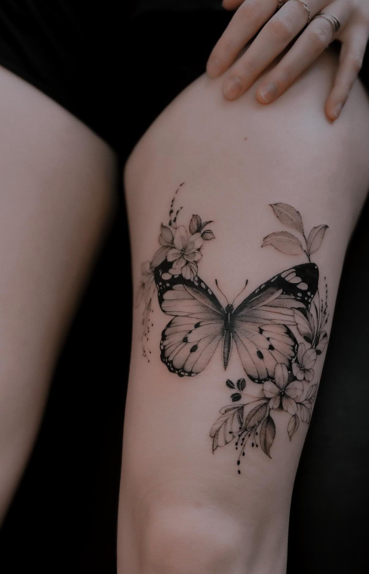 Butterfly tattoo surrounded by wildflowers and leaves on a person's thigh