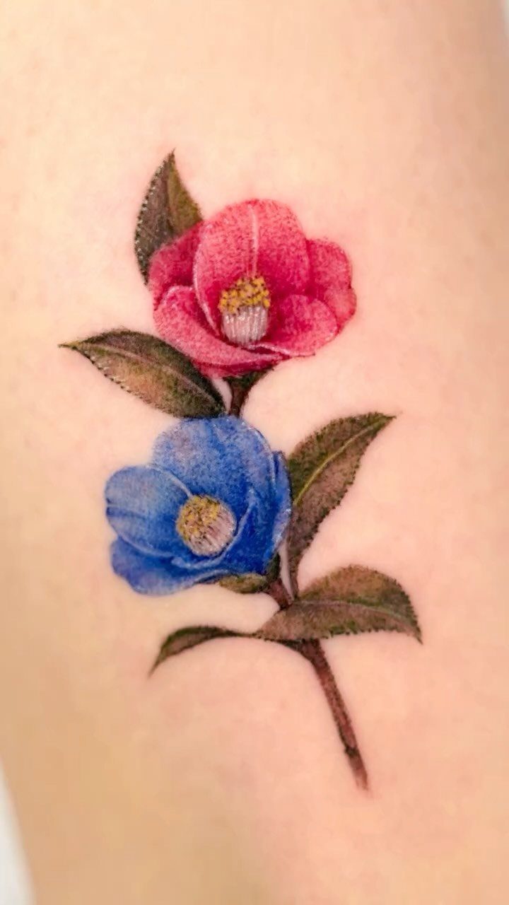 Two camellia flowers, one red and one blue, tattooed on skin with leaves.