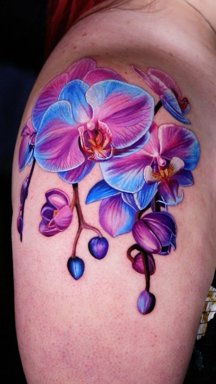Orchid flower tattoo in purple and blue hues on a person's shoulder.