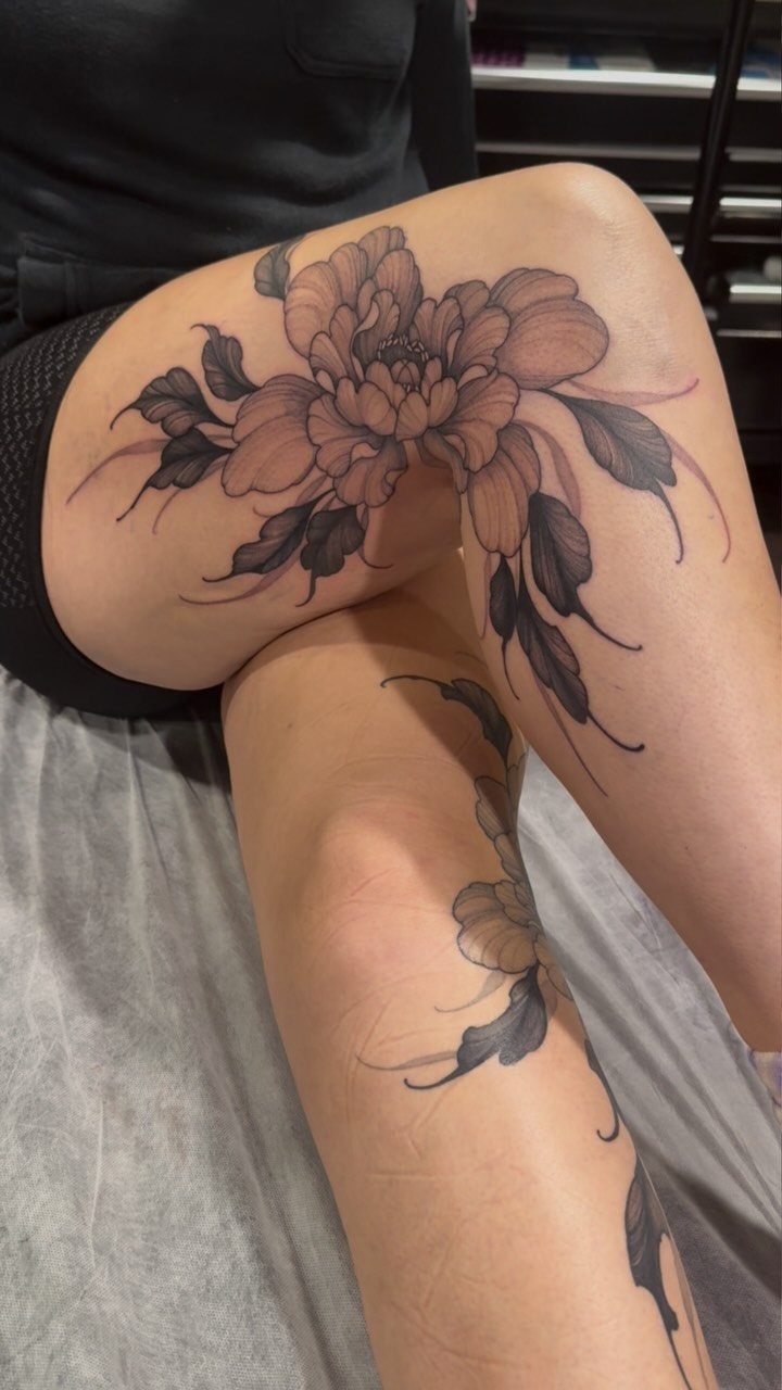 Large peony flower tattoo with soft pink petals and dark leaves on a thigh.