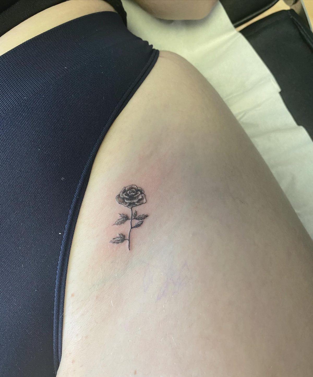 Small black ink rose tattoo with stem and leaves on a person's hip