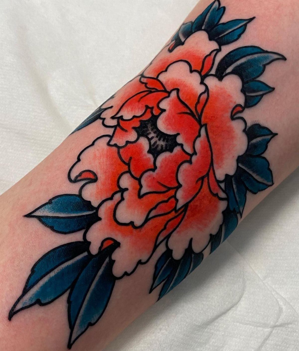 Peony flower tattoo with orange petals and dark blue leaves on an arm