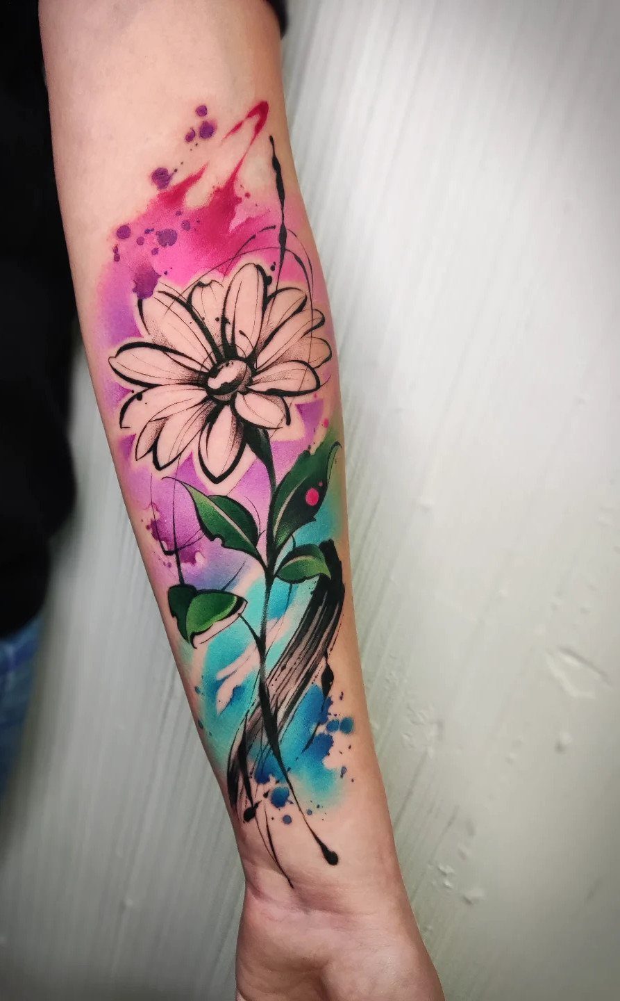 Watercolor flower tattoo on a forearm with pink and blue ink splashes.