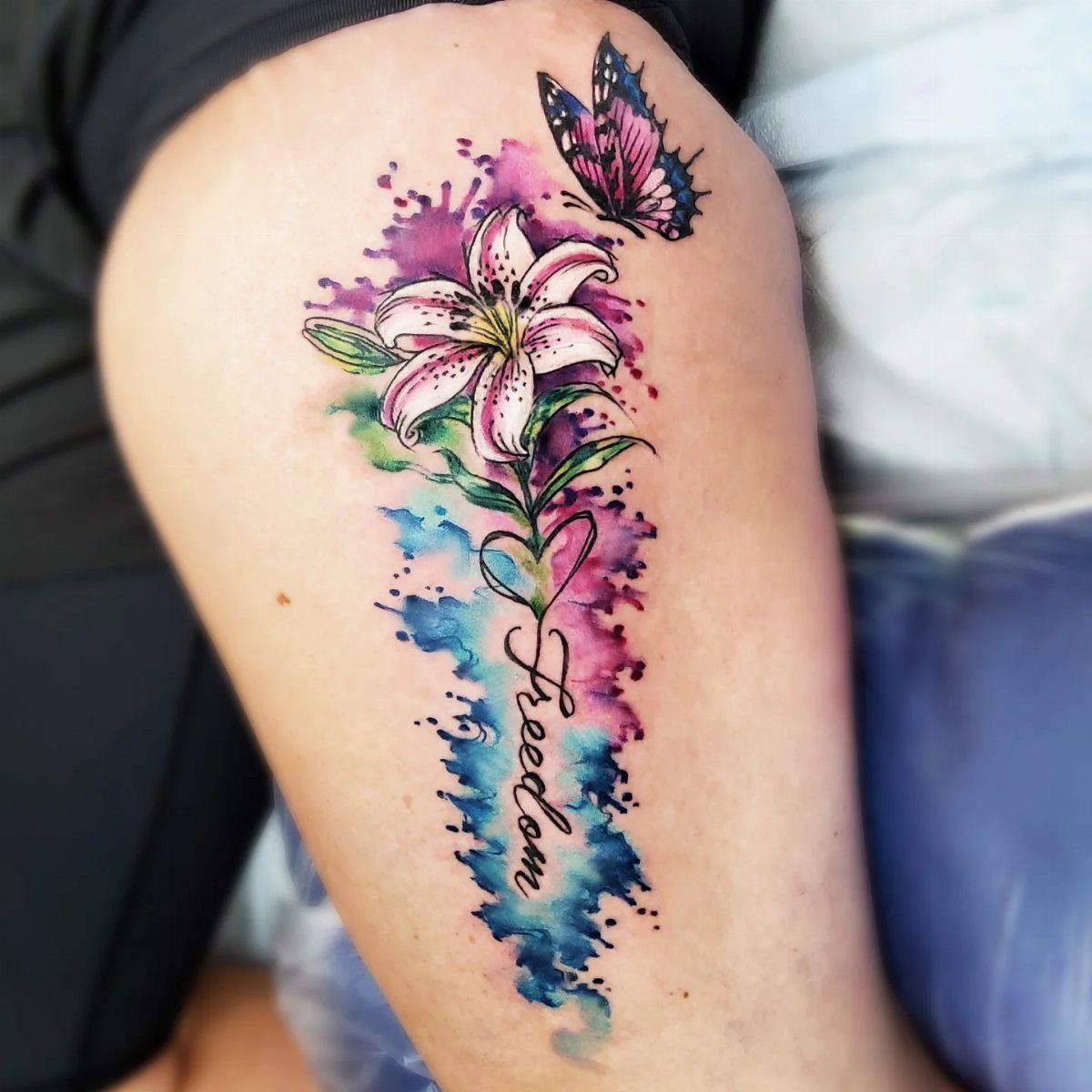 Watercolor lily tattoo with a butterfly and the word Freedom on a leg