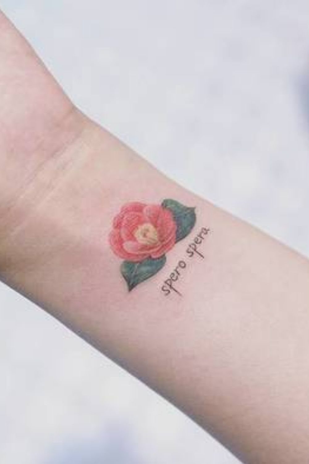 Pink camellia flower tattoo with green leaves and cursive text on a wrist