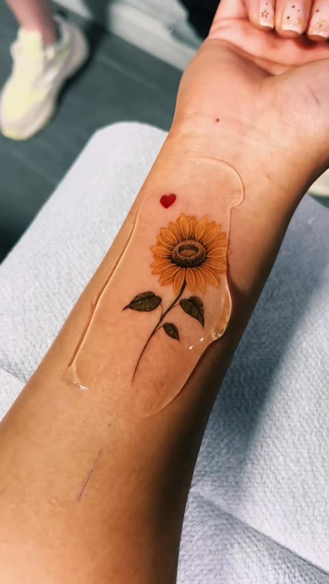 Sunflower tattoo with a small red heart on a person's inner wrist