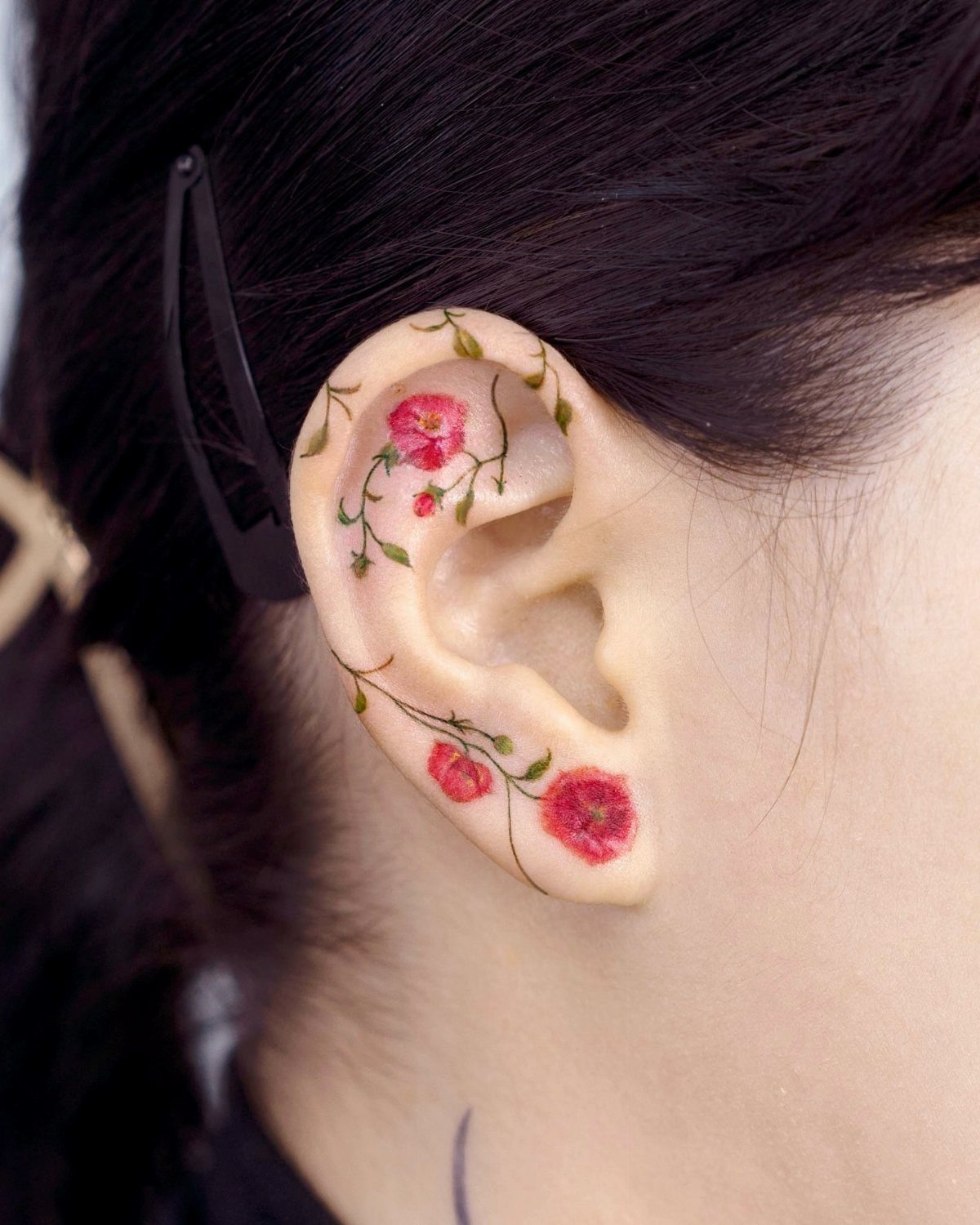 Small red flower and green vine tattoo curving around a person's ear.