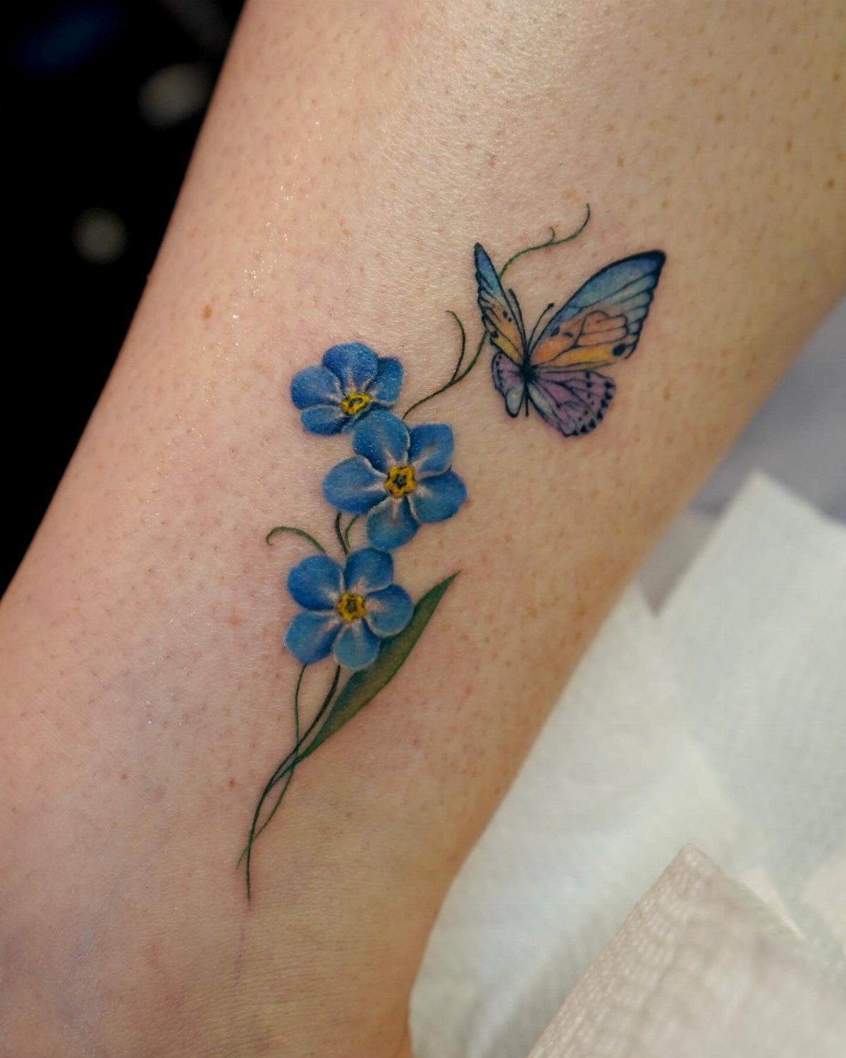 Blue forget-me-not flower tattoo with a small butterfly on a person's skin
