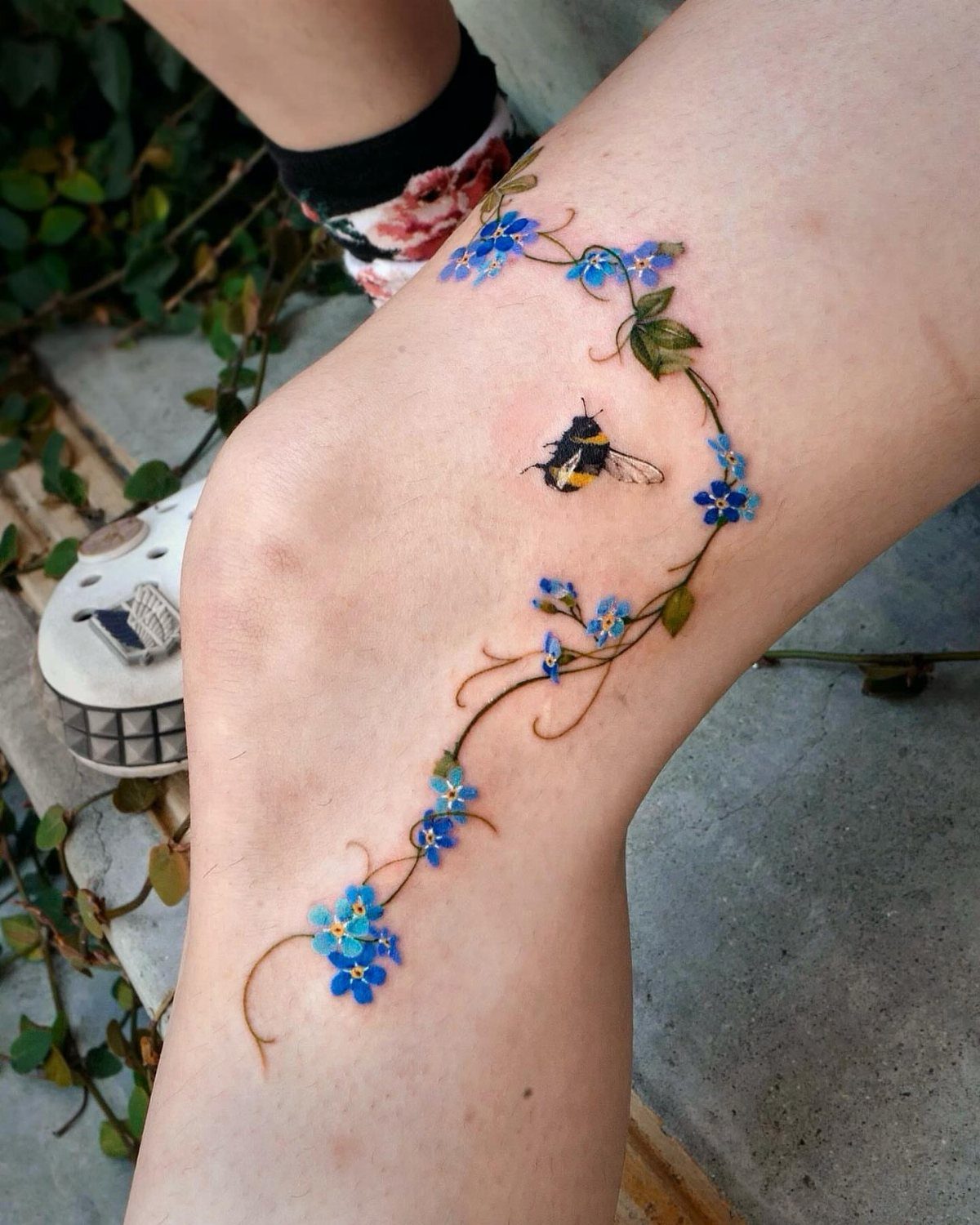 Colorful forget-me-not tattoo with a bumblebee on a person's knee and leg.