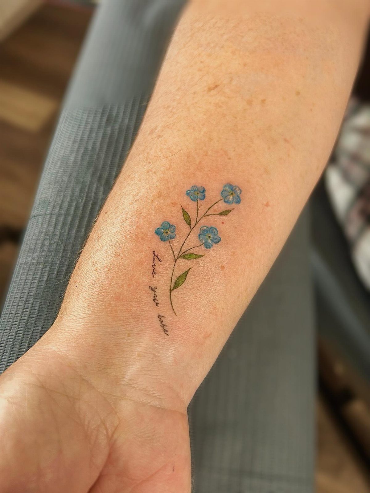 Blue forget-me-not flower tattoo with green leaves and cursive text on a forearm