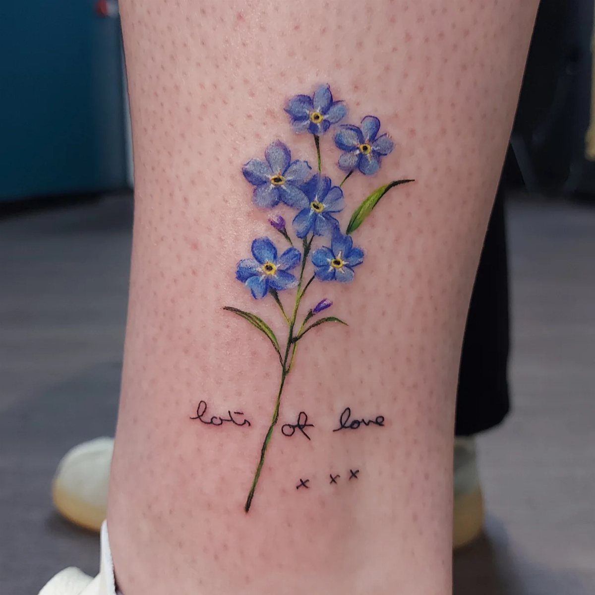 Blue forget-me-not flower tattoo with green leaves and text on a leg