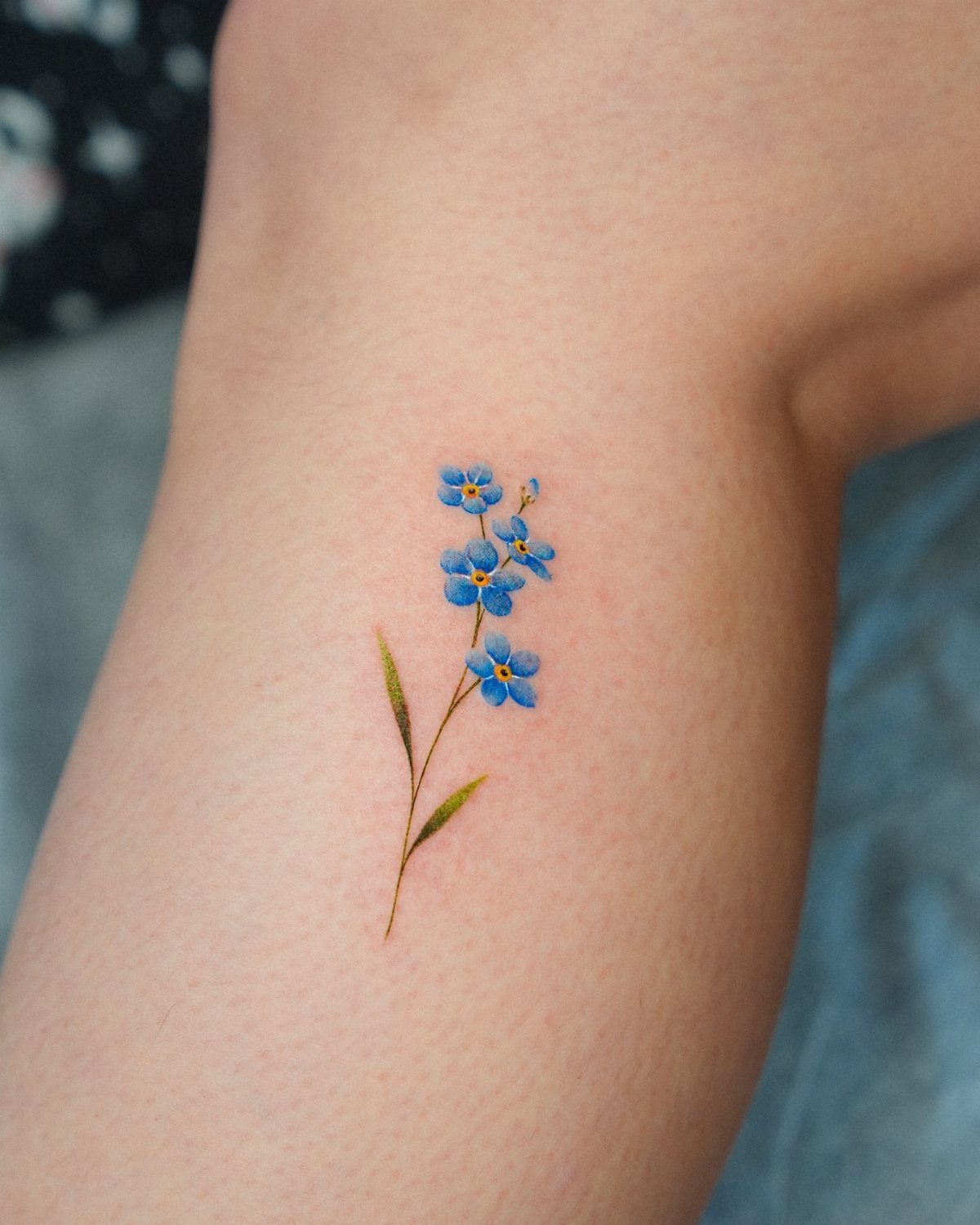 Small blue forget-me-not flower tattoo with green leaves on a person's leg.