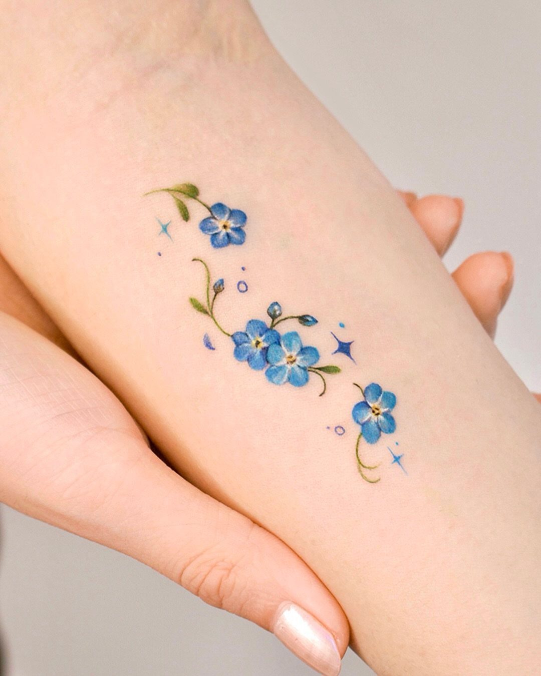 Small blue forget-me-not flower tattoo with green leaves on a person's forearm.