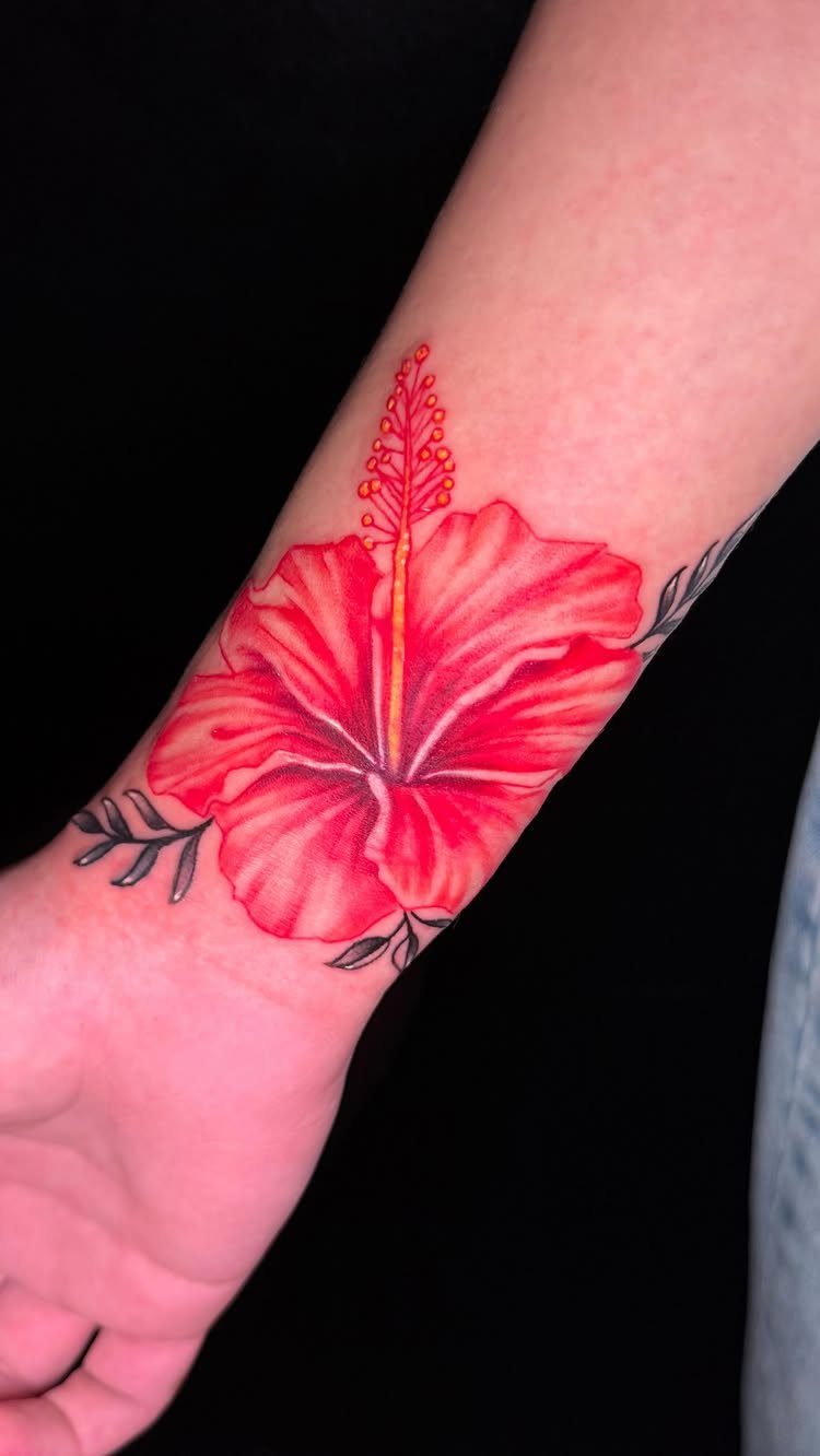 Red hibiscus flower tattoo with yellow stamen on a person's inner wrist.
