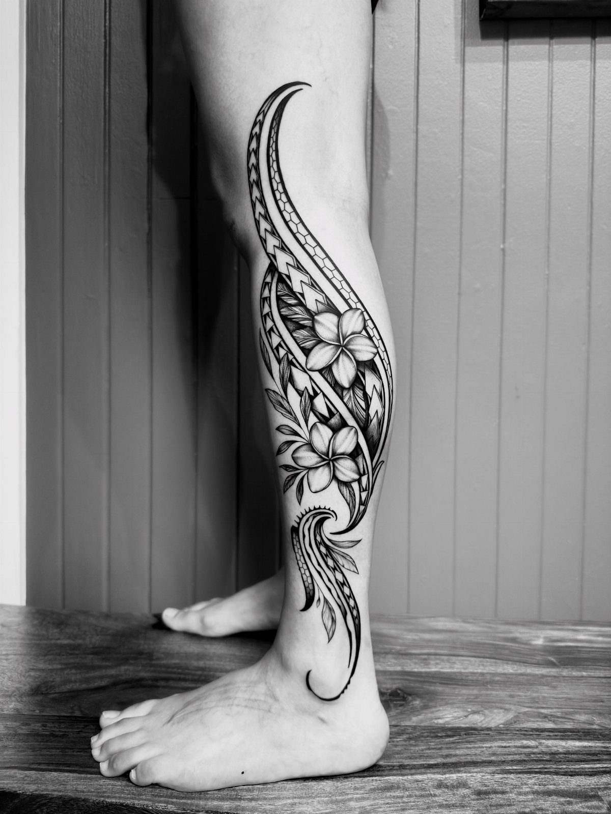 Black and gray tropical flower tattoo with tribal patterns on a lower leg.