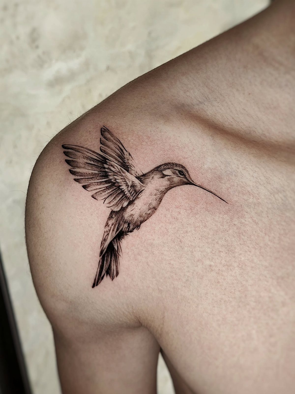 Black ink hummingbird tattoo in flight on a person's shoulder with detailed shading.