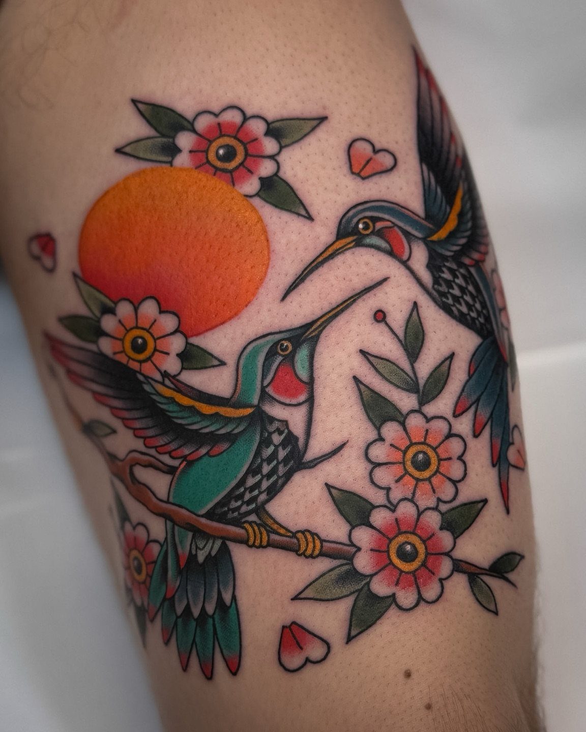 Traditional tattoo of two colorful hummingbirds with pink flowers and an orange sun.