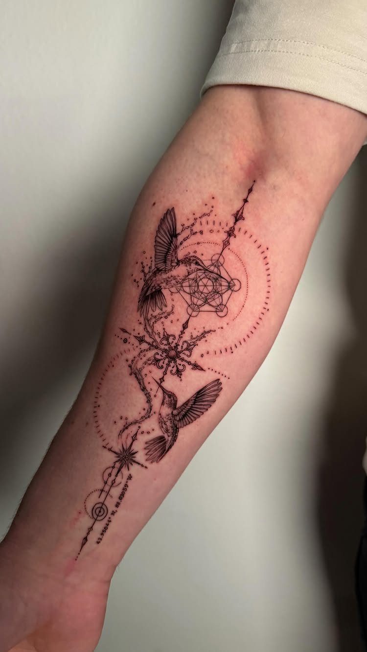 Two hummingbird tattoos on a forearm with a central geometric design and dots.