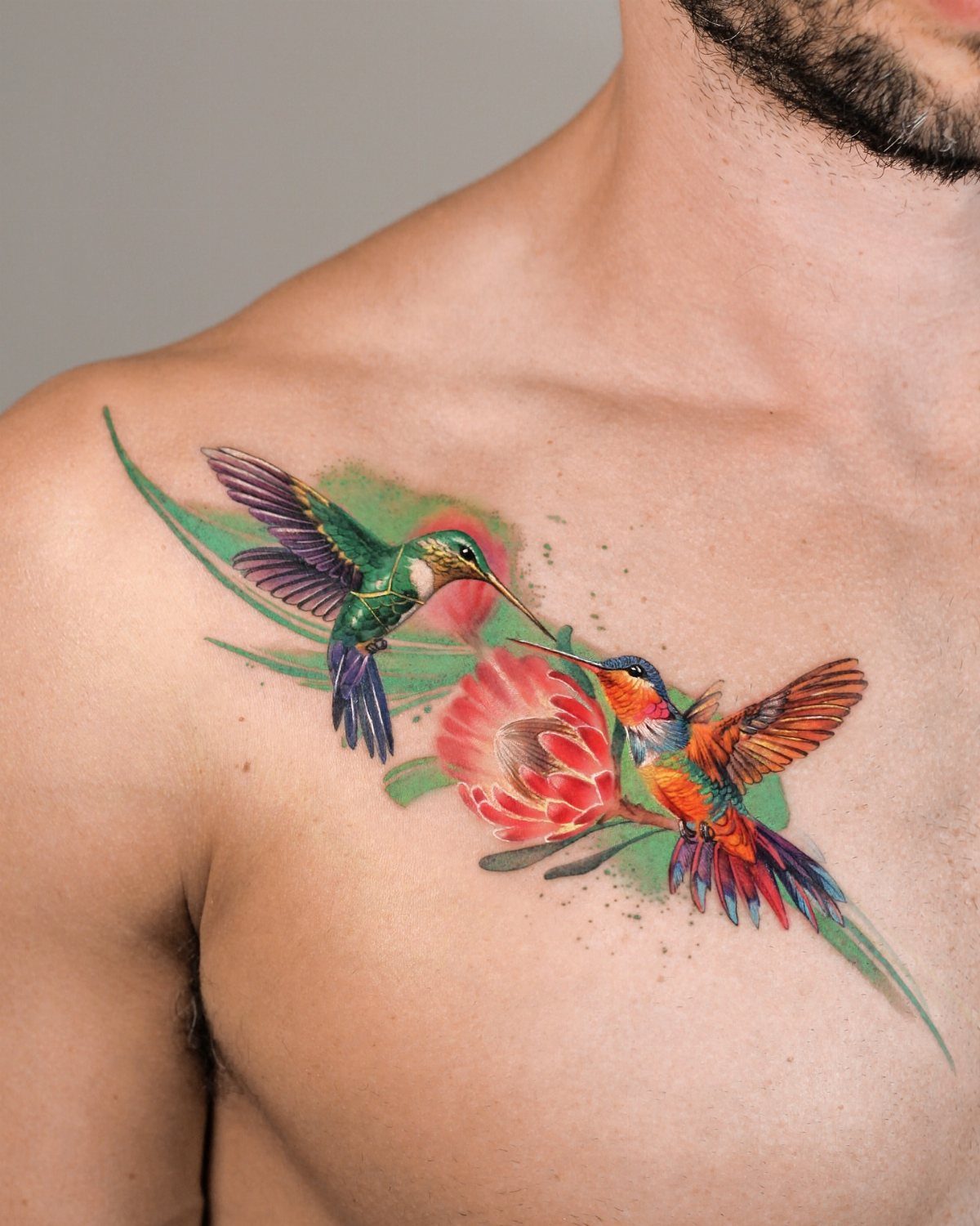 Two colorful hummingbird tattoos facing each other with a red flower on a chest.