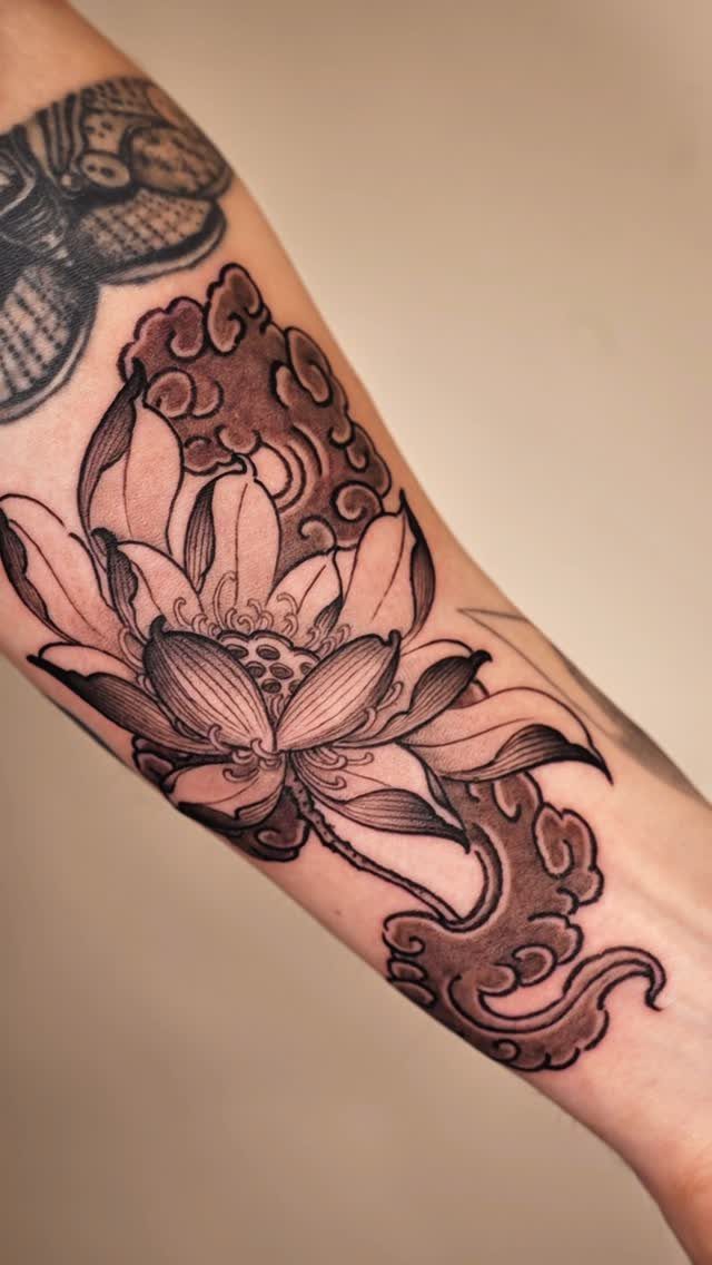 Lotus flower tattoo with stylized clouds on a person's forearm in black and grey.