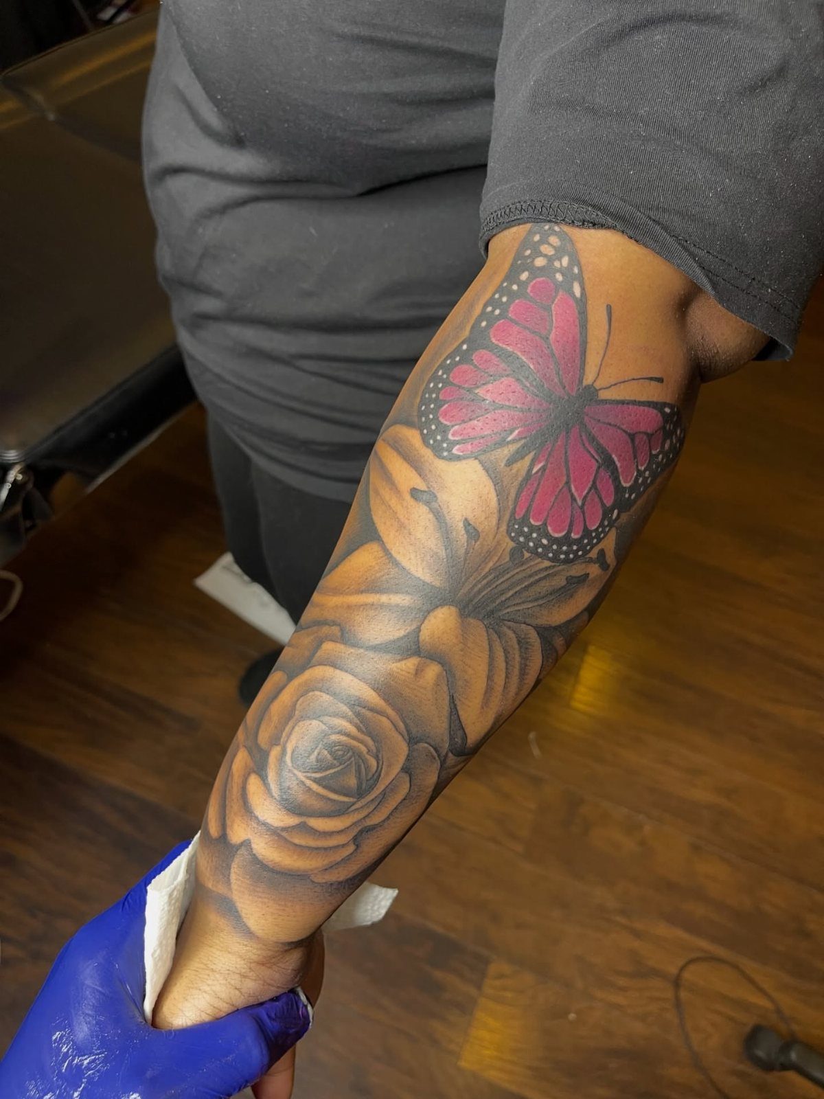 Forearm tattoo featuring a pink butterfly, a lily, and a rose in detail.