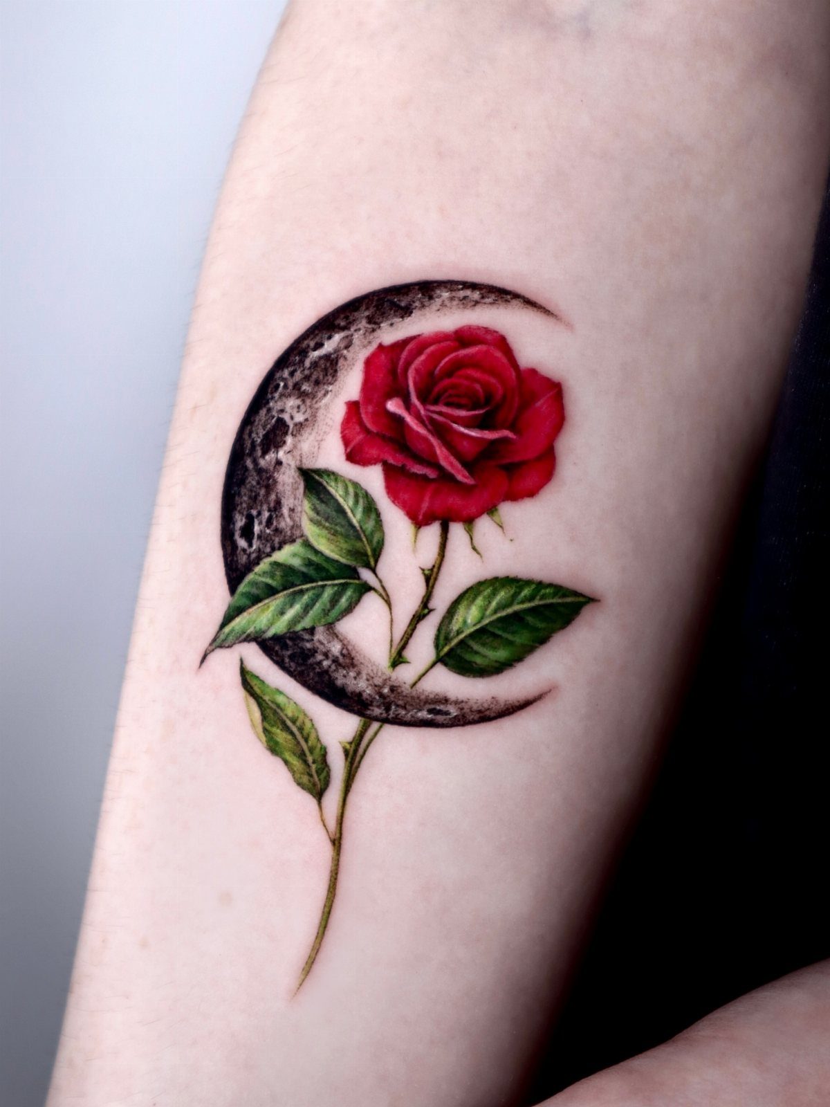 Red rose tattoo with green leaves and a dark crescent moon on forearm