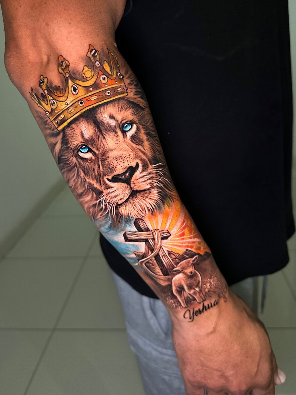 Lion wearing a golden crown tattoo on a man's forearm with religious imagery.