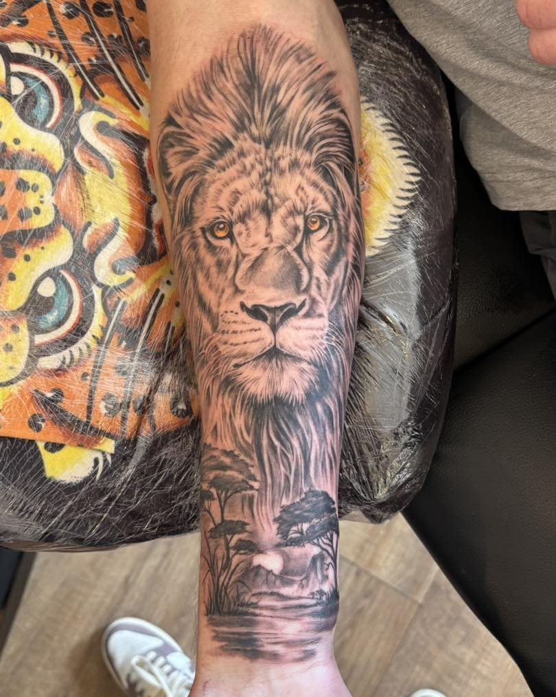 Detailed black and grey lion tattoo on a forearm with a landscape base.